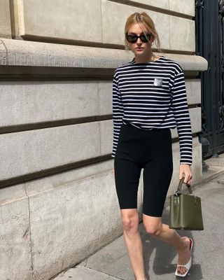 fashion influencer Camille Charriere walking on a sidewalk in France wearing black sunglasses, a striped long sleeve t-shirt, capri bike shorts, green top-handle bag, and white square-toe mule heel sandals