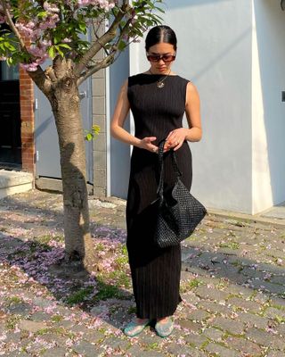 fashion influencer Sasha Mei poses next to a tree with pink flowers wearing black oval sunglasses, a sleeveless black plissé maxi dress, cord necklace, woven bag, and blue jelly sandals