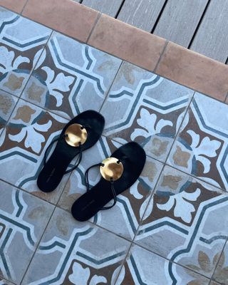 black flat Tory Burch sandals with a metal medallion on a printed tile floor shot by fashion editor Anna Laplaca