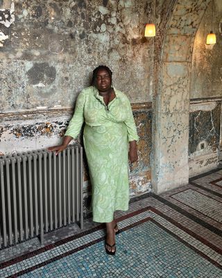 British fashion influencer Abisola Omole poses in front of a distressed wall and mosaic tile floors wearing a light green wrap dress and black flip flop heeled sandals